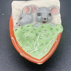 May include: A ceramic trinket dish in the shape of a shield, featuring two gray mice nestled on a green bed. The dish has a red border and a white pillow. The mice have pink ears and red mouths.