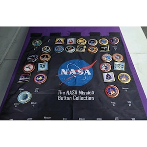 May include: A collection of NASA mission buttons displayed on a black fabric with a purple border. The buttons feature various designs and logos, including the NASA logo and mission names. The text "The NASA Mission Button Collection" is visible.