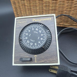 May include: A square Sears Programmer electrical timer with a wood-grain finish faceplate, a black circular dial featuring AM, PM, NOON, and NIGHT markings, and a black push-button switch labeled PUSH and OFF, connected to a black power cord.