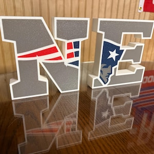May include: Two freestanding block letters N and E in a silver glitter finish with white borders. The letter N features a red and blue striped graphic, while the letter E displays a blue and white profile of a person wearing a hat.