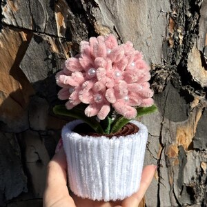 May include: A handmade pink flower arrangement with pearl accents in a white corduroy pot. The flower has a fluffy texture and green leaves. The pot is held against a textured brown background.