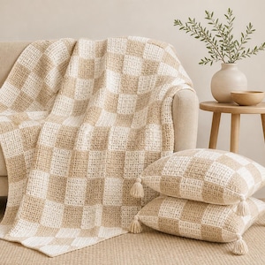 May include: A beige and cream checkered crochet throw blanket draped over a beige sofa. Two matching square cushions with tassels are stacked on the floor. A small wooden side table holds a vase with greenery and a wooden bowl.