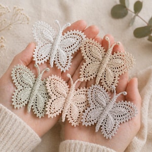 May include: Six handmade, crocheted butterfly appliques in pastel shades of white, beige, and pale green. The butterflies are held in open hands against a soft, neutral background, highlighting their delicate, lace-like details. Each butterfly is unique.