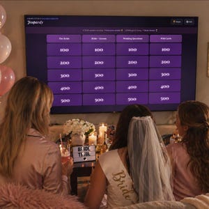 May include: A group of women gather for a bachelorette party, watching a Jeopardy game on a large screen. The game board is purple with white text. Balloons and a sign that says "Bride Tribe" are visible.