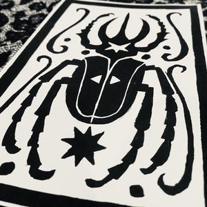 May include: A black and white rectangular print featuring a stylized beetle illustration centered within a thick black border. The beetle design includes a star on its head, two triangular markings on its back, and a prominent eight-pointed star below.