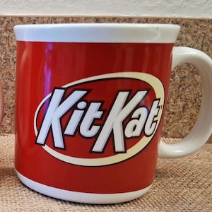 May include: Two red ceramic mugs with white rims and handles. Each mug features the Kit Kat logo in white letters on a yellow and white oval. The mugs are on a brown surface.