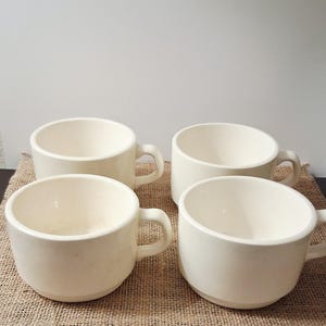 May include: Four off-white ceramic mugs with handles. The mugs are arranged on a burlap surface. The mugs have a simple, rounded design, suitable for coffee, tea, or other beverages. The neutral color and classic shape make them versatile for any kitchen.