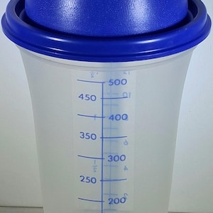 May include: A clear plastic shaker with a blue lid and a blue rim. The shaker has a conical shape and features blue measurement markings in milliliters, ranging from 50 ml to 500 ml. The lid has a textured surface.