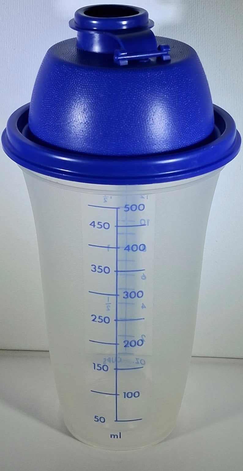 May include: A clear plastic shaker with a blue lid and a blue rim. The shaker has a conical shape and features blue measurement markings in milliliters, ranging from 50 ml to 500 ml. The lid has a textured surface.