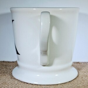 May include: A white ceramic mug with a curved handle. The mug has a wide base and a slightly flared top. A black letter is visible on the side of the mug. The mug is set against a neutral background.