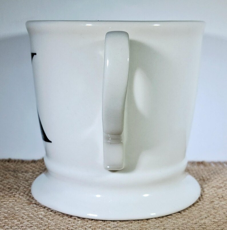 May include: A white ceramic mug with a curved handle. The mug has a wide base and a slightly flared top. A black letter is visible on the side of the mug. The mug is set against a neutral background.