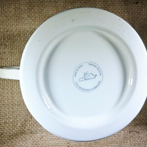 May include: White ceramic mug with a handle, viewed from the bottom. The base has a circular design with a bird illustration and the text "MADE IN CHINA". The mug is dishwasher and microwave safe.