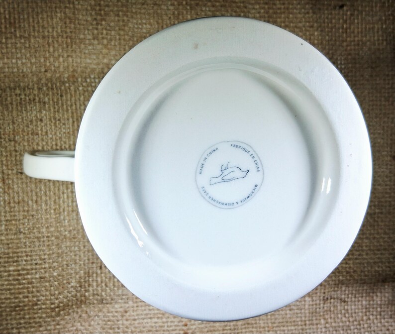 May include: White ceramic mug with a handle, viewed from the bottom. The base has a circular design with a bird illustration and the text "MADE IN CHINA". The mug is dishwasher and microwave safe.