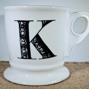 May include: White ceramic mug featuring a large black letter "K" with an ornate design. The mug has a rounded base and a curved handle. The design is detailed and decorative.