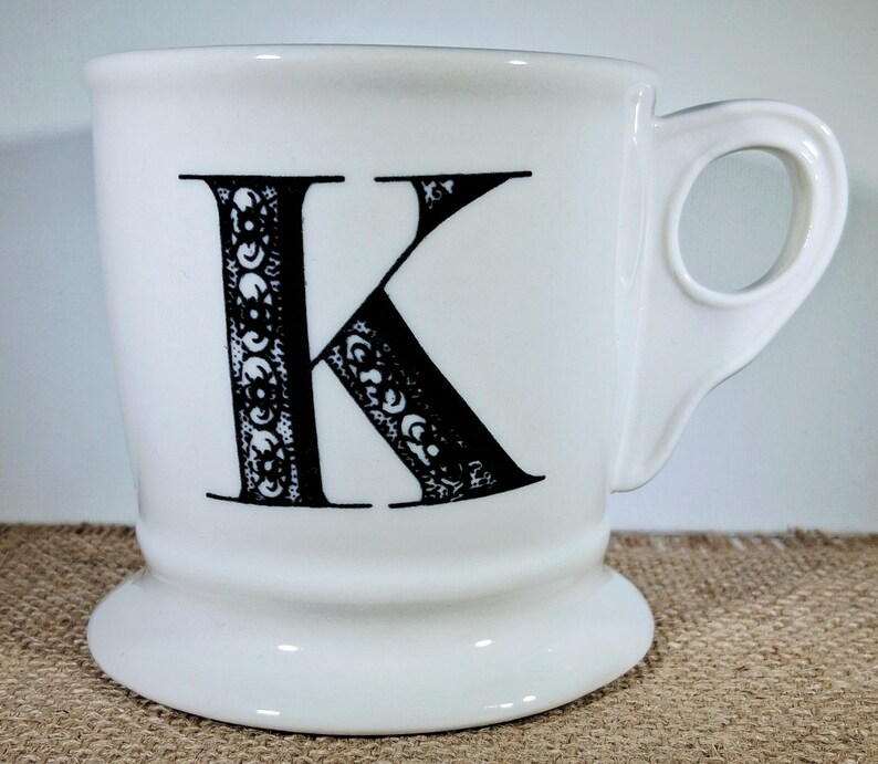 May include: White ceramic mug featuring a large black letter "K" with an ornate design. The mug has a rounded base and a curved handle. The design is detailed and decorative.