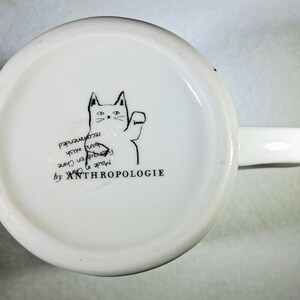 Puede incluir: Taza de cer&aacute;mica blanca con asa, con una ilustraci&oacute;n negra de un gato saludando. El texto "ANTHROPOLOGIE" est&aacute; impreso debajo del gato. La parte inferior de la taza tambi&eacute;n incluye texto que indica que est&aacute; hecha en China y se recomienda lavar a mano.