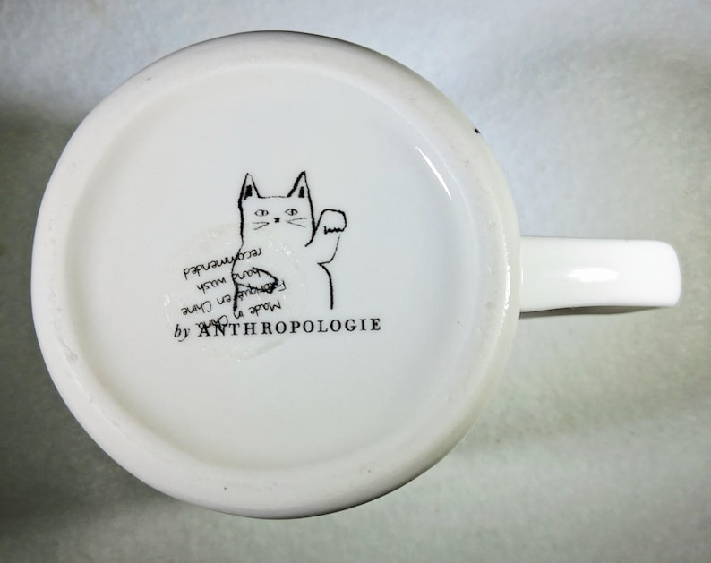 Puede incluir: Taza de cer&aacute;mica blanca con asa, con una ilustraci&oacute;n negra de un gato saludando. El texto "ANTHROPOLOGIE" est&aacute; impreso debajo del gato. La parte inferior de la taza tambi&eacute;n incluye texto que indica que est&aacute; hecha en China y se recomienda lavar a mano.