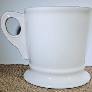 May include: A white ceramic mug with a rounded handle and a pedestal base. The mug has a smooth, glossy finish and a simple, classic design. The mug is suitable for coffee, tea, or other beverages.
