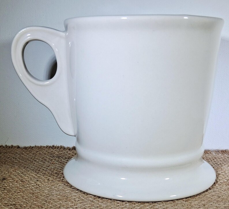 May include: A white ceramic mug with a rounded handle and a pedestal base. The mug has a smooth, glossy finish and a simple, classic design. The mug is suitable for coffee, tea, or other beverages.
