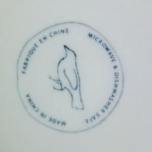 May include: A white ceramic surface with a blue circular stamp. The stamp features a bird perched on a branch, with text that reads "FABRIQU&Eacute; EN CHINE", "MICROWAVE & DISHWASHER SAFE", and "MADE IN CHINA".