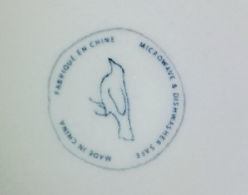 May include: A white ceramic surface with a blue circular stamp. The stamp features a bird perched on a branch, with text that reads "FABRIQU&Eacute; EN CHINE", "MICROWAVE & DISHWASHER SAFE", and "MADE IN CHINA".