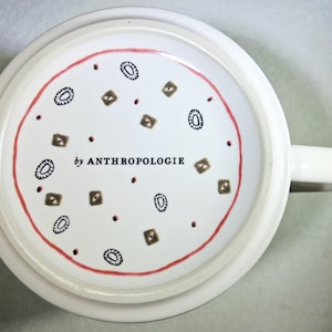 May include: White ceramic mug with a handle, featuring a red circular border and a pattern of brown and black geometric shapes and dots. The text "by ANTHROPOLOGIE" is printed in black.