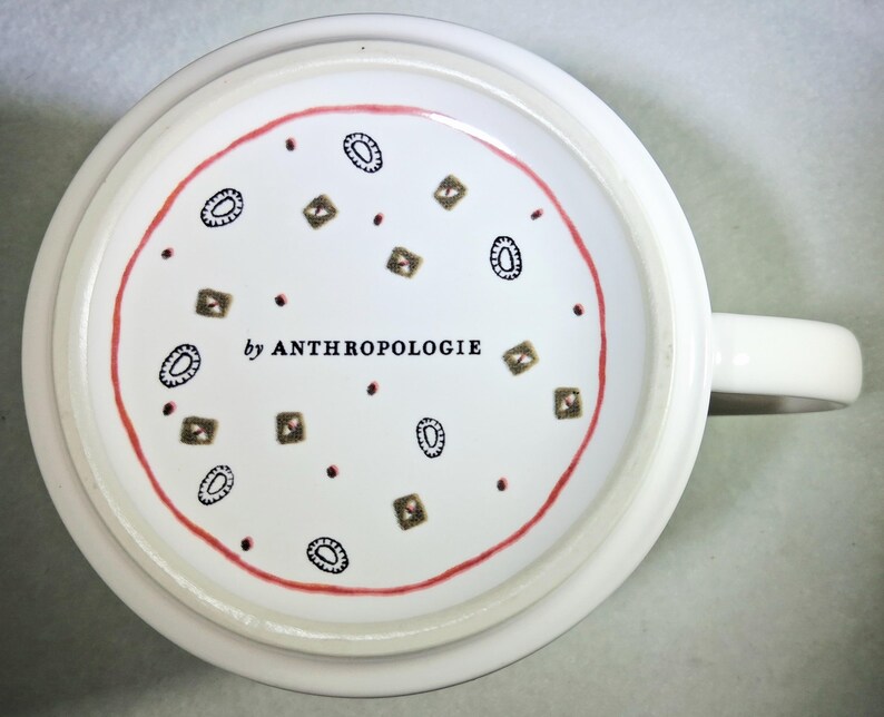 May include: White ceramic mug with a handle, featuring a red circular border and a pattern of brown and black geometric shapes and dots. The text "by ANTHROPOLOGIE" is printed in black.