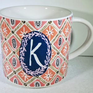 May include: White ceramic mug with a decorative pattern in coral, pink, and gold. A navy blue oval with the letter "K" in white is centered on the mug. The mug has a curved handle.
