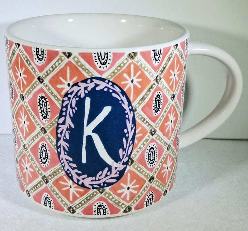 May include: White ceramic mug with a decorative pattern in coral, pink, and gold. A navy blue oval with the letter "K" in white is centered on the mug. The mug has a curved handle.