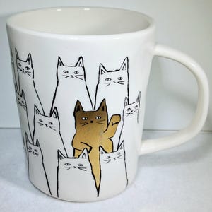 Puede incluir: Taza de cer&aacute;mica blanca con un dise&ntilde;o caprichoso de gatos. La taza presenta un patr&oacute;n repetido de caras de gatos en blanco y negro, con un gato marr&oacute;n saludando. La taza tiene un asa curvada.
