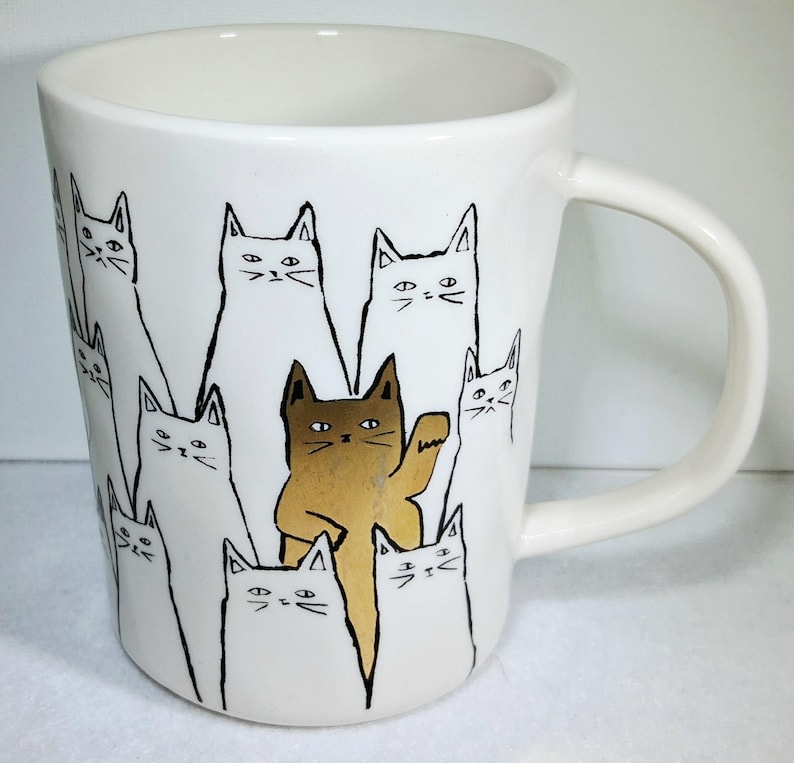Puede incluir: Taza de cer&aacute;mica blanca con un dise&ntilde;o caprichoso de gatos. La taza presenta un patr&oacute;n repetido de caras de gatos en blanco y negro, con un gato marr&oacute;n saludando. La taza tiene un asa curvada.