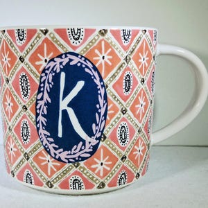 May include: White ceramic mug with a decorative pattern in coral, pink, and gold. The mug features a blue oval with a white letter "K" and a floral border. The handle is white.