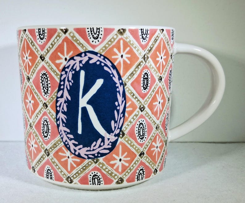 May include: White ceramic mug with a decorative pattern in coral, pink, and gold. The mug features a blue oval with a white letter "K" and a floral border. The handle is white.