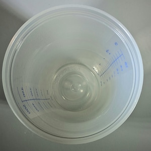 May include: A clear plastic measuring cup with blue measurement markings. The cup has a wide, circular opening and a slightly textured interior. The measurements are in milliliters, ranging from 50 to 400.