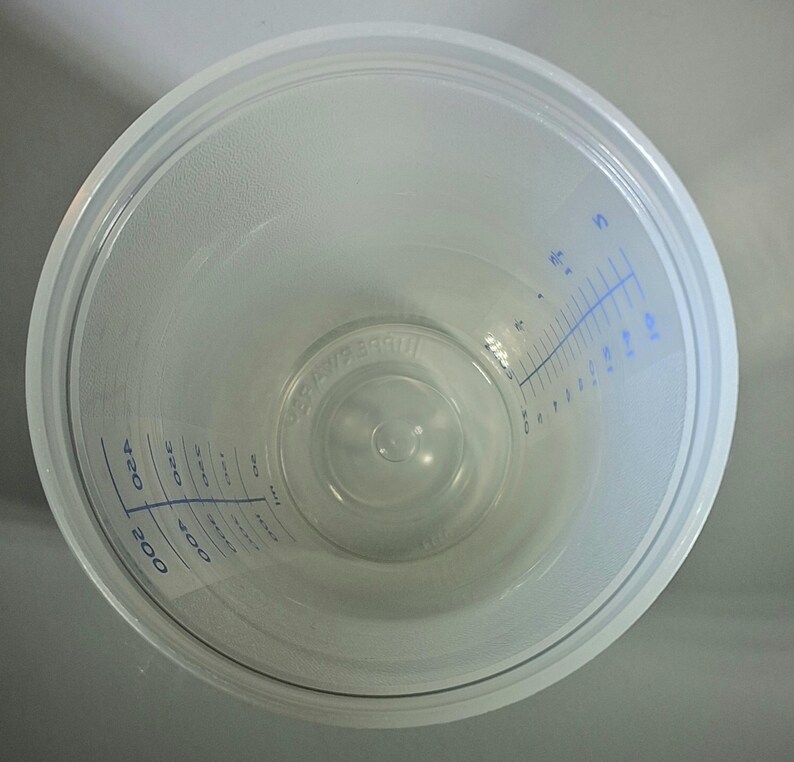 May include: A clear plastic measuring cup with blue measurement markings. The cup has a wide, circular opening and a slightly textured interior. The measurements are in milliliters, ranging from 50 to 400.