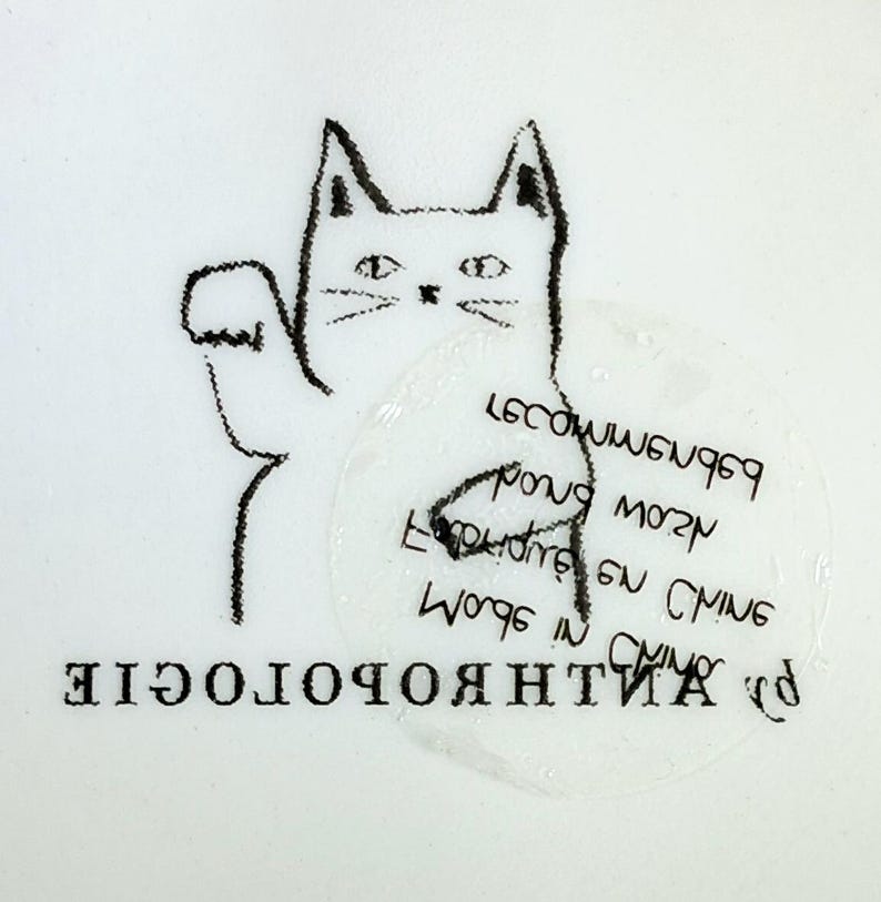 Puede incluir: Ilustraci&oacute;n en blanco y negro de un gato con la pata levantada. El gato est&aacute; dibujado con un estilo sencillo y esbozado. La palabra "ANTHROPOLOGIE" est&aacute; impresa en la parte inferior, y hay texto en un c&iacute;rculo cerca del gato.