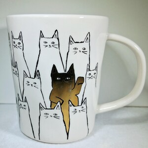 Puede incluir: Taza de cer&aacute;mica blanca con un dise&ntilde;o de gatos negros y dorados. El gato central es dorado y levanta una pata, rodeado de gatos blancos con contornos negros. La taza tiene un asa curva.