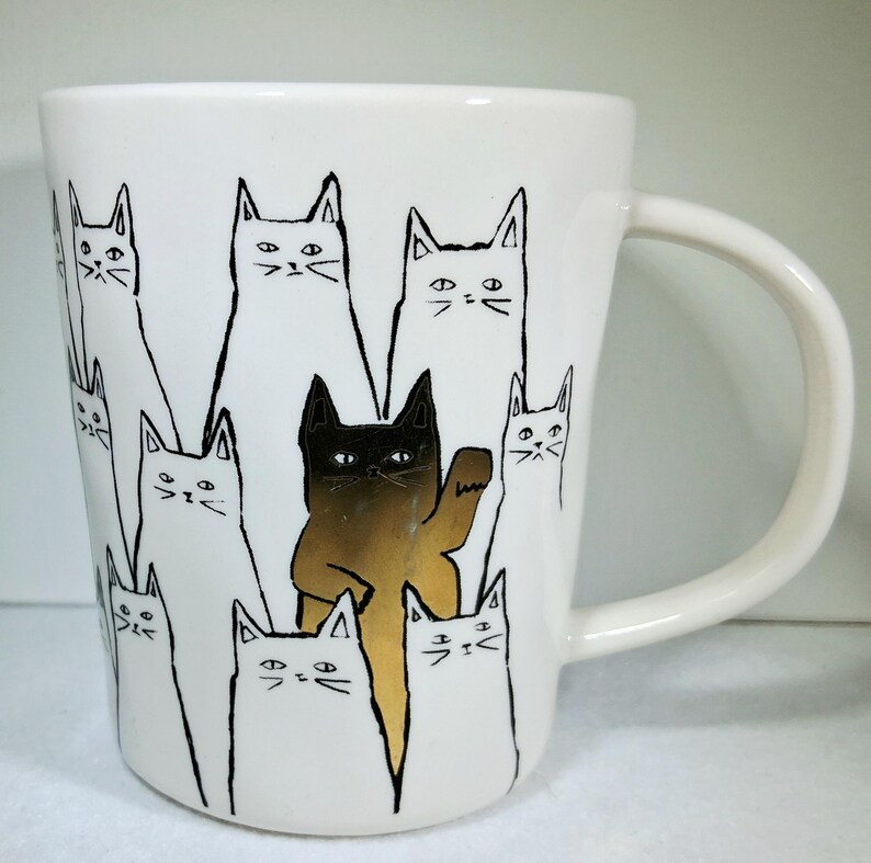 Puede incluir: Taza de cer&aacute;mica blanca con un dise&ntilde;o de gatos negros y dorados. El gato central es dorado y levanta una pata, rodeado de gatos blancos con contornos negros. La taza tiene un asa curva.