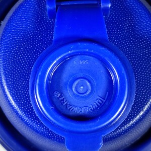 May include: Close-up of a blue Tupperware lid with a textured surface. The lid features a hinged tab and the brand name "Tupperware" is visible. The number "564-3" is printed on the lid.
