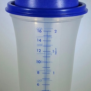 May include: A clear plastic shaker with a blue lid and cap. The shaker has measurement markings in ounces and cups, ranging from 2 to 16 ounces. The blue lid has a textured surface.