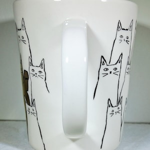 Puede incluir: Taza de cer&aacute;mica blanca con asa, decorada con dibujos lineales negros de gatos estilizados. Los gatos tienen orejas triangulares, rasgos faciales simples y cuerpos largos. La taza es de tama&ntilde;o est&aacute;ndar.