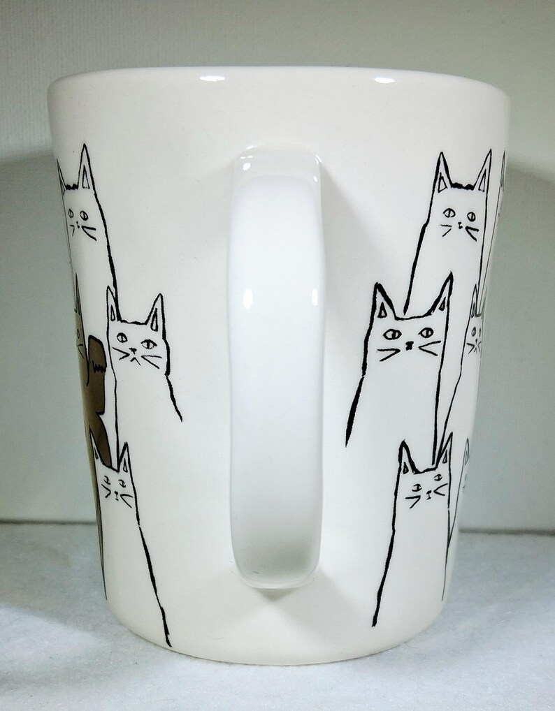 Puede incluir: Taza de cer&aacute;mica blanca con asa, decorada con dibujos lineales negros de gatos estilizados. Los gatos tienen orejas triangulares, rasgos faciales simples y cuerpos largos. La taza es de tama&ntilde;o est&aacute;ndar.