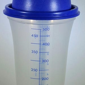 May include: A clear plastic shaker with a blue lid and a blue measuring scale in milliliters. The shaker has a capacity of 500 ml, with markings at 50 ml increments. The lid has a textured surface.
