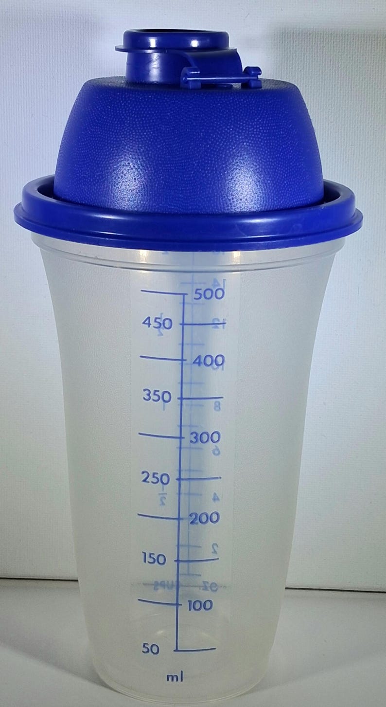 May include: A clear plastic shaker with a blue lid and a blue measuring scale in milliliters. The shaker has a capacity of 500 ml, with markings at 50 ml increments. The lid has a textured surface.