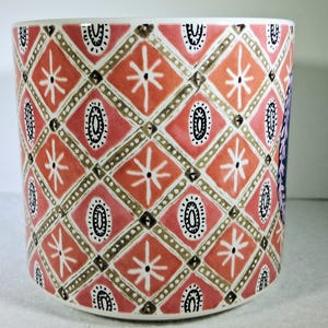 May include: A cylindrical ceramic planter with a repeating geometric pattern. The design features diamond shapes in shades of coral and pink, with white starbursts and black oval accents. The planter has a white interior and base.