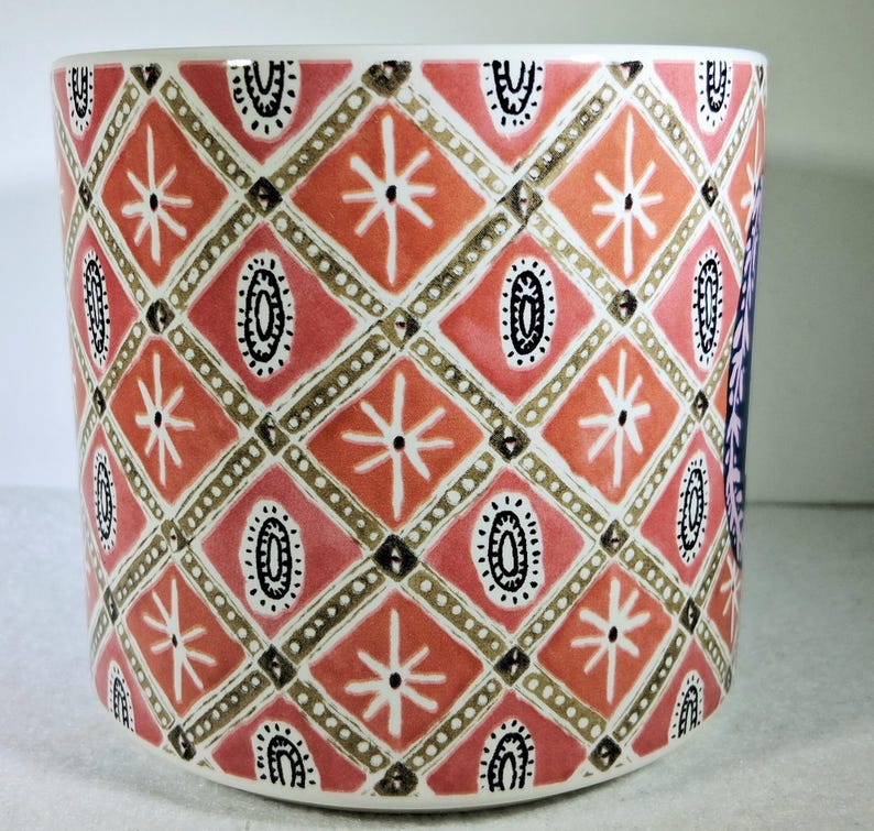 May include: A cylindrical ceramic planter with a repeating geometric pattern. The design features diamond shapes in shades of coral and pink, with white starbursts and black oval accents. The planter has a white interior and base.