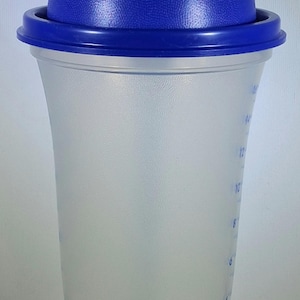 May include: A clear plastic shaker bottle with a blue lid and cap. The bottle has measurement markings in ounces and a curved shape. The lid has a textured surface and a flip-top closure.