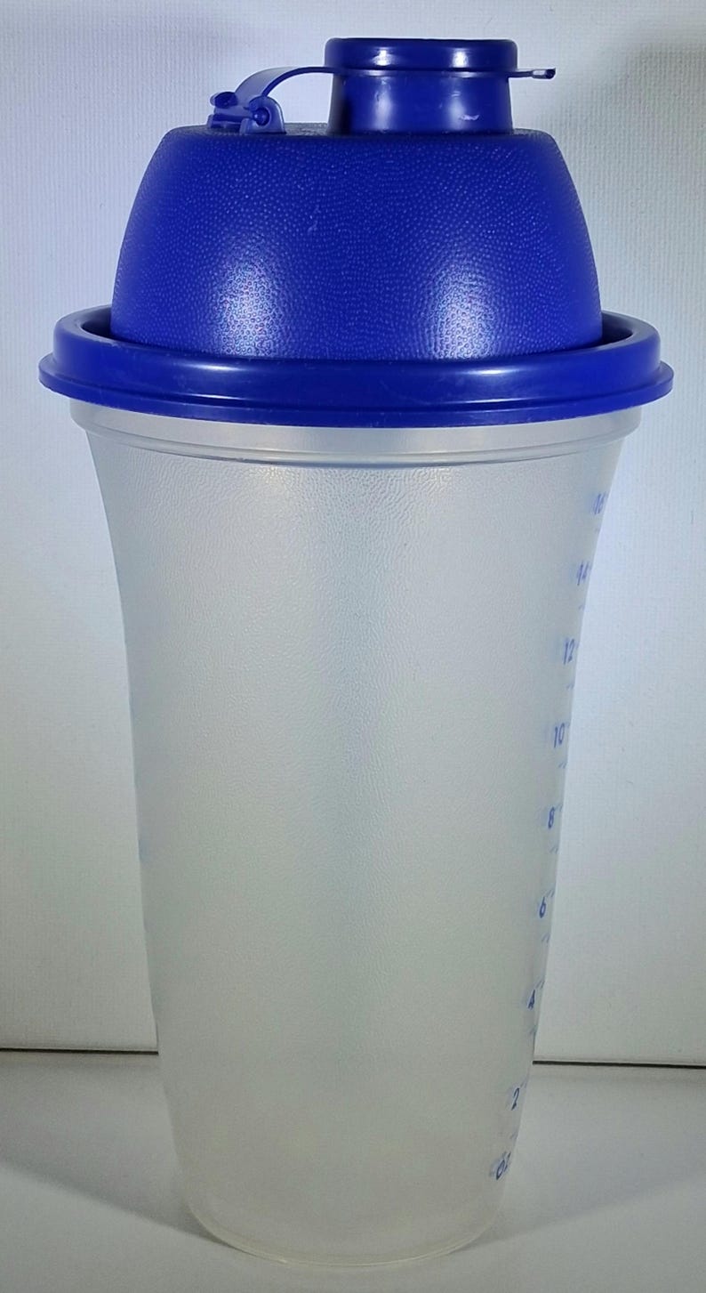 May include: A clear plastic shaker bottle with a blue lid and cap. The bottle has measurement markings in ounces and a curved shape. The lid has a textured surface and a flip-top closure.