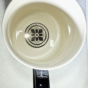 以下が含まれることがあります： 黒いハンドルが付いた白いセラミックマグ。内側には、底に黒と白の花柄モザイクのデザインが施されています。マグカップは光沢のある仕上がりです。