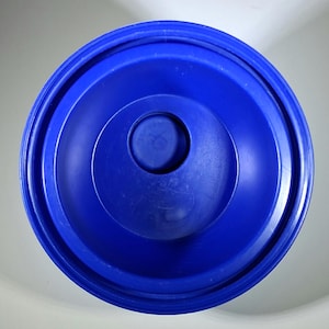 May include: A top-down view of a solid blue plastic lid. The lid has a circular design with concentric rings and a raised center. The plastic appears to be smooth and glossy, with a slight sheen. The lid is likely for a container.